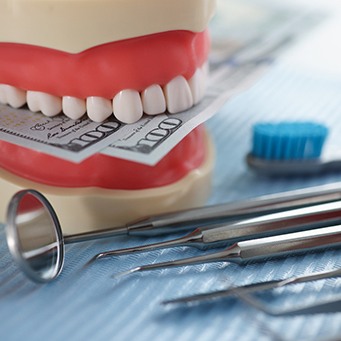 A jaw mockup biting hundred-dollar bills next to some dental tools