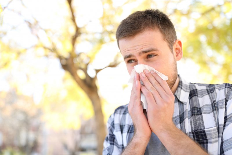 a man wearing a gray plaid shirt uses a tissue to blow his nose