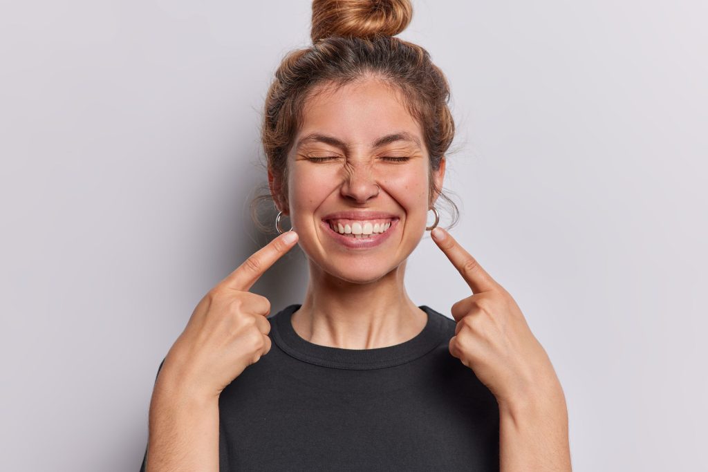 Woman with hair in bun squinting eyes shut and pointing at her smile with both hands
