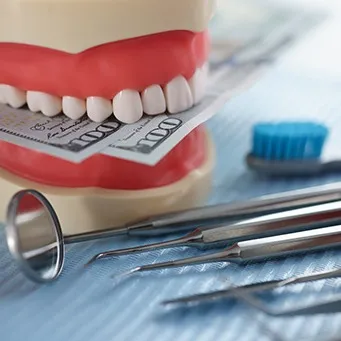 A jaw mockup biting hundred-dollar bills next to some dental tools