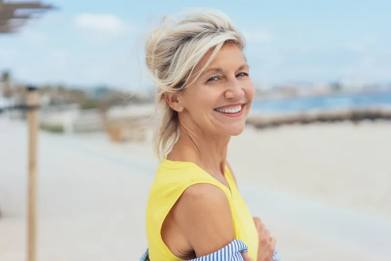 an older woman wearing a yellow blouse overlooks the beach while showing off her dental implants in Vero Beach