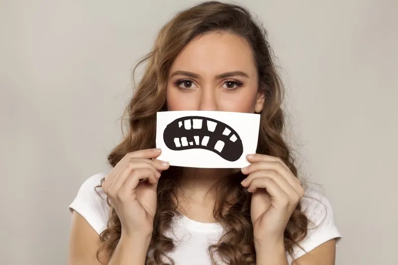 A woman holding an illustration of a poor smile over her mouth.